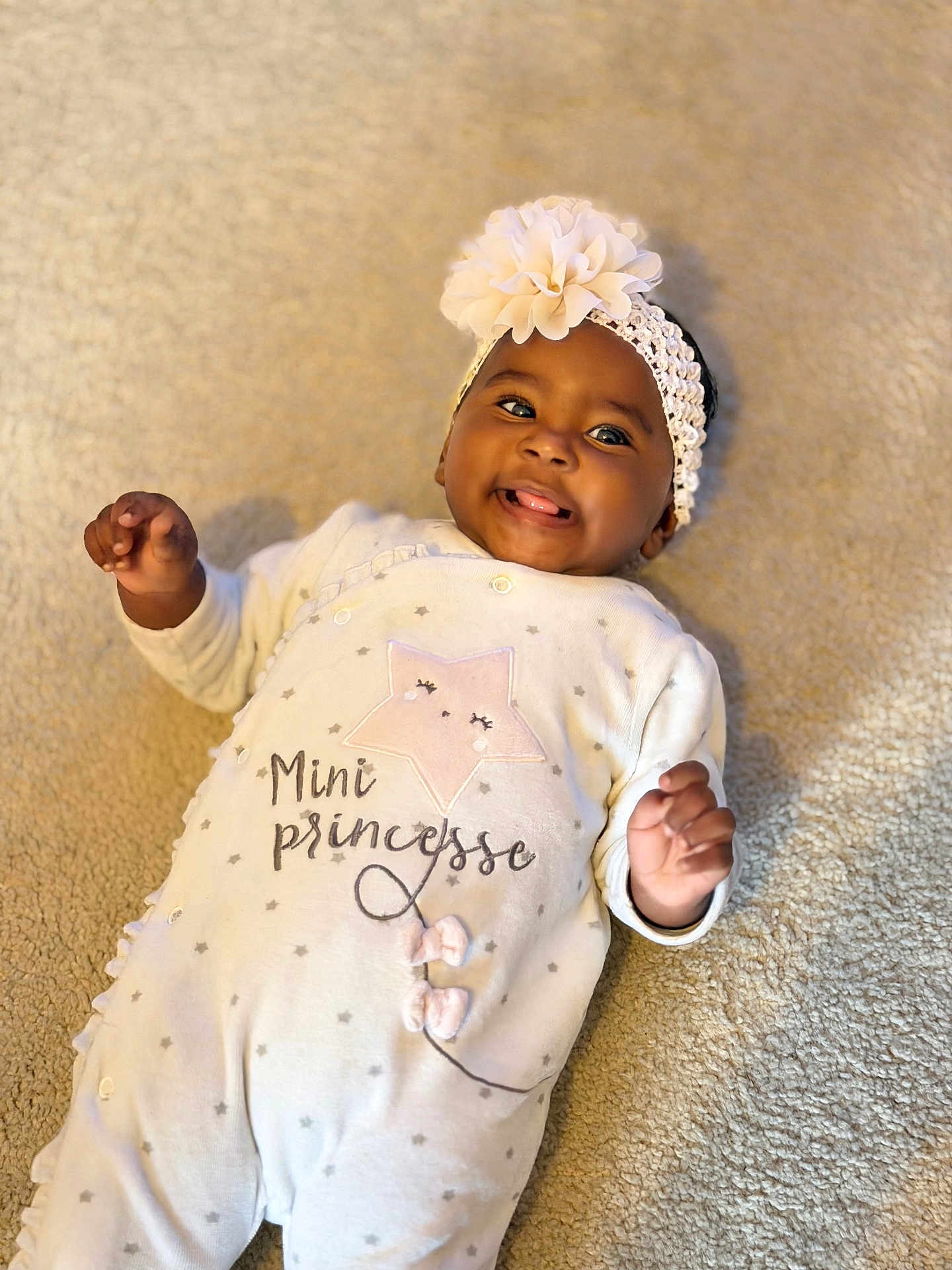Naïma a rejoint le concours — aidez-le/la à gagner de superbes lots ! baby, infant, smiling, onesie, headband, flower, carpet, lying_down, happy, cute, child, portrait, indoor, soft_texture, clothing, face, hands, skin, expression, playful