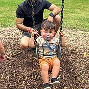 Isaac joined the competition — help win amazing prizes! child, toddler, pacifier, swing, playground, adult, man, grass, wood_chips, outdoor, park, casual_clothing, shorts, tshirt, sneakers, smiling, family, fun, daylight, recreation