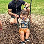 child, toddler, pacifier, swing, playground, adult, man, grass, wood_chips, outdoor, park, casual_clothing, shorts, tshirt, sneakers, smiling, family, fun, daylight, recreation