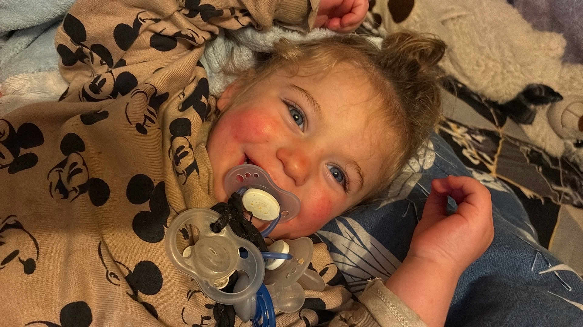 Mylan a rejoint le concours — aidez-le/la à gagner de superbes lots ! child, toddler, baby, pacifier, pacifiers, smile, blue_eyes, rosy_cheeks, blanket, mickey_mouse_print, pajamas, bed, stuffed_animal, hair_bun, hand, closeup, portrait, cozy, playful, interior