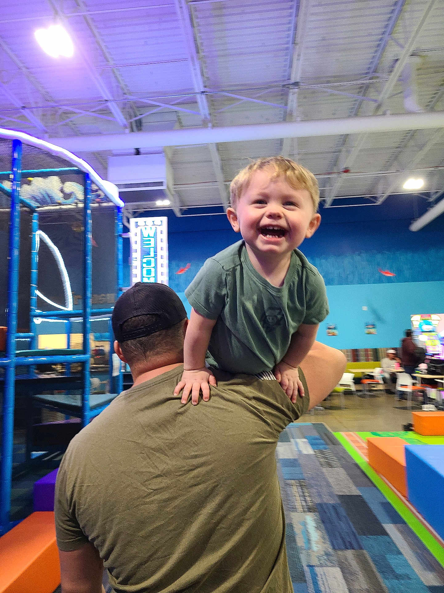 Colton joined the competition — help win amazing prizes! toddler, child, adult, indoor, play_area, smiling, happy, shoulder_ride, cap, green_shirt, carpet, colorful, playground_equipment, netting, people, chairs, tables, blue_wall, lighting, fun