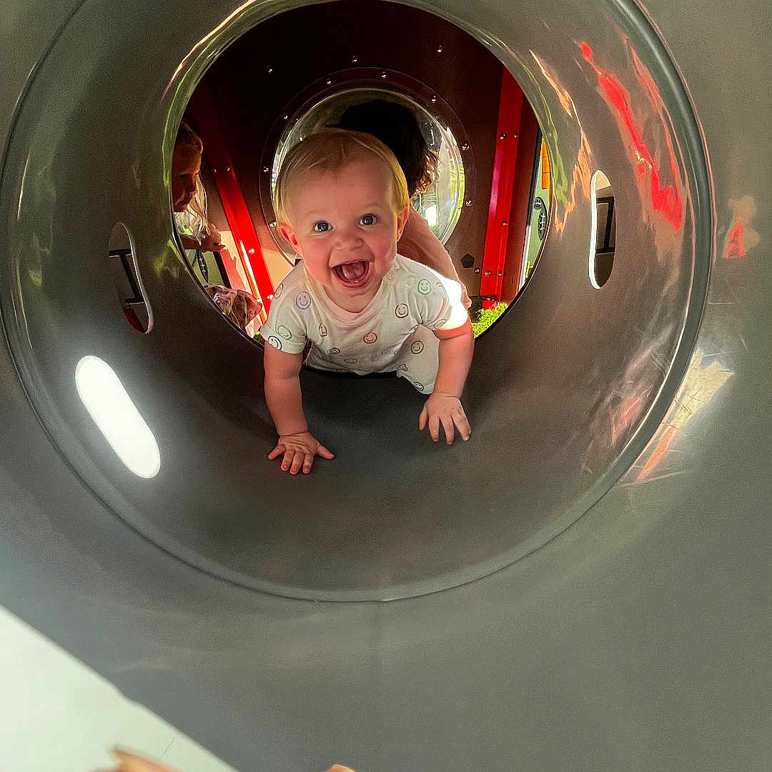 Emerson is registered to the contest to win money with this photo: baby, child, playground, tunnel, crawling, smiling, happy, person, outdoor, fun, joy, play, curious, hand, finger, clothing, light, reflection, face, toy