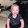 Emerson is registered to the contest to win money with this photo: baby, smiling, indoor, child, puma_shirt, black_clothing, sitting, happy, face, blue_eyes, person, infant, cute, young_child, home, television, blanket, floor, comfort, portrait
