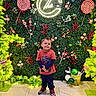 child, boy, smiling, standing, red_shirt, blue_pants, sneakers, holiday_decor, candy, lollipops, greenery, ornaments, floor_tiles, indoor, festive, bright_colors, decorations, neon_sign, happy, portrait