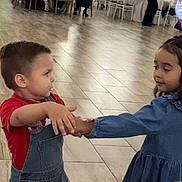 child, boy, girl, indoor, dance, holding_hands, overalls, dress, floor, tile_floor, event, people, table, chair, party, celebration, flooring, interaction, clothing, happy