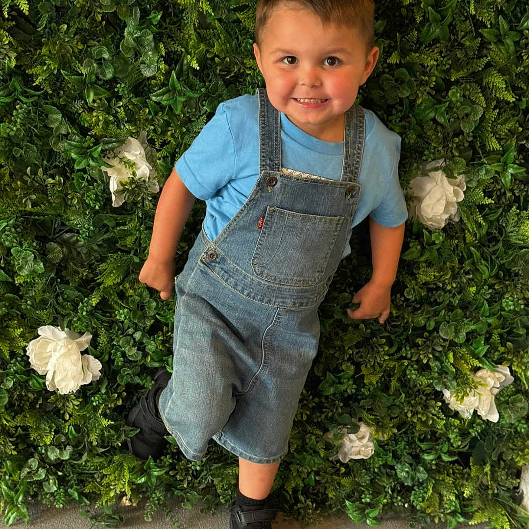 Xander joined the competition — help win amazing prizes! toddler, child, smiling, denim_overalls, blue_shirt, greenery, leafy_wall, white_flowers, standing, casual_clothing, happy, cute, portrait, indoor, plants, nature_background, black_shoes, short_hair, playful, youth