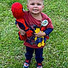 child, boy, smiling, toy, spiderman, plush_toy, birthday_badge, red_shirt, blue_pants, sneakers, grass, outdoor, happy, cute, portrait, person, standing, playful, celebration, daylight