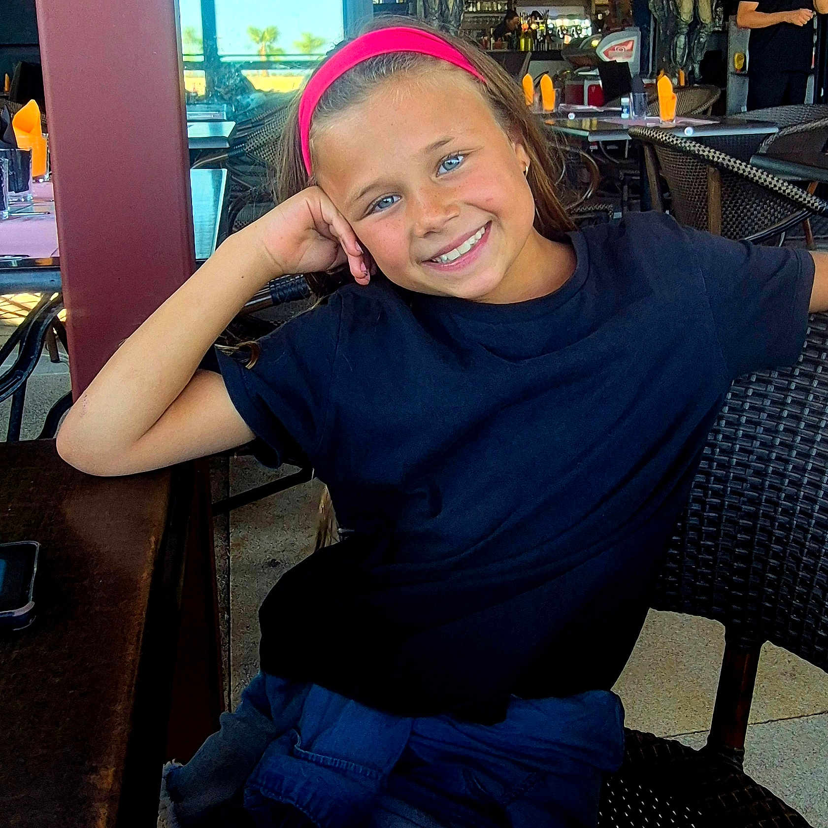 Lélina participe au concours pour gagner de l'argent avec cette photo : black_shirt, blue_eyes, casual, chair, child, dining, girl, happy, headband, indoor, people, person, portrait, relaxed, restaurant, seated, smiling, table, wicker_chair, young