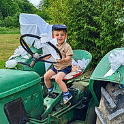 Armand a rejoint le concours — aidez-le/la à gagner de superbes lots ! child, toddler, tractor, steering_wheel, cap, shorts, shoes, white_bow, greenery, outdoor, grass, seat_cushion, rustic, vintage, smiling, nature, sunny, vehicle, playful, happy
