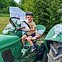 child, toddler, tractor, steering_wheel, cap, shorts, shoes, white_bow, greenery, outdoor, grass, seat_cushion, rustic, vintage, smiling, nature, sunny, vehicle, playful, happy