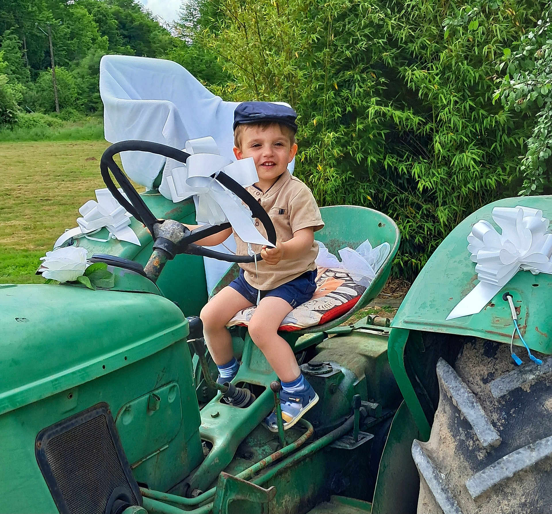 Armand a rejoint le concours — aidez-le/la à gagner de superbes lots ! child, toddler, tractor, steering_wheel, cap, shorts, shoes, white_bow, greenery, outdoor, grass, seat_cushion, rustic, vintage, smiling, nature, sunny, vehicle, playful, happy