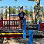 child, playground, toddler, outdoor, tree, fence, bus, sunlight, shadow, wood, clothing, person, smile, blue, red, yellow, pants, sneakers, daytime, sky