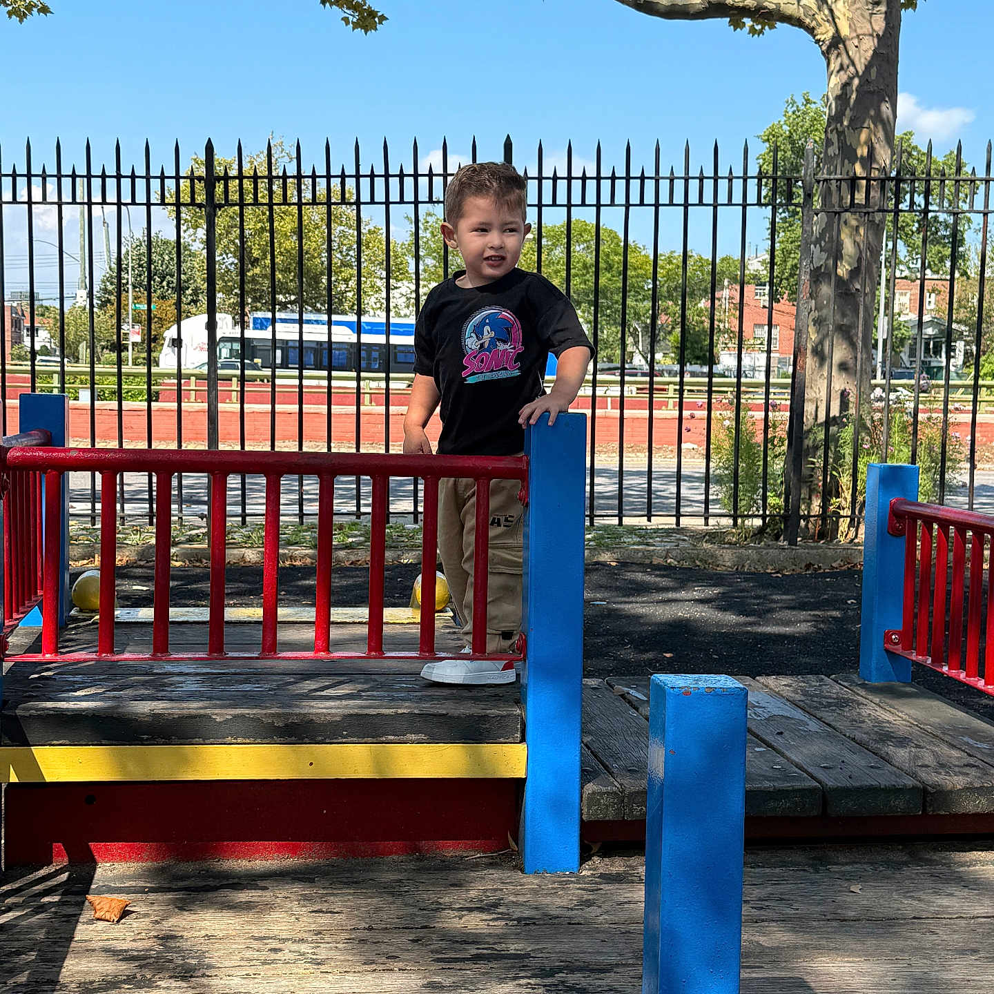 Cesar Nahuel is registered to the contest to win money with this photo: blue, bus, child, clothing, daytime, fence, outdoor, pants, person, playground, red, shadow, sky, smile, sneakers, sunlight, toddler, tree, wood, yellow