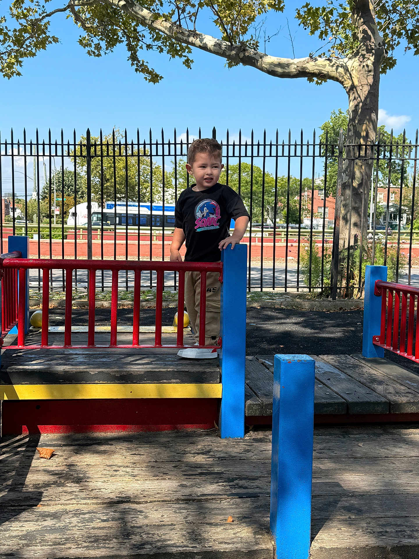 Cesar Nahuel is registered to the contest to win money with this photo: child, playground, toddler, outdoor, tree, fence, bus, sunlight, shadow, wood, clothing, person, smile, blue, red, yellow, pants, sneakers, daytime, sky