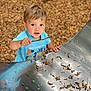 activity, blue_shirt, casual_clothing, child, curious, daylight, exploration, fun, kid, metal_slide, nature, outdoor, park, person, playground, smiling, stick, toddler, wood_chips, young_child