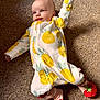baby, infant, carpet, onesie, pineapple_pattern, footwear, slippers, strawberry_design, lying_down, raised_arm, cute, child, indoors, soft_lighting, smiling_face, small_person, cozy_clothing, floor, playful, adorable