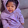 Aylah is registered to the contest to win money with this photo: baby, child, clothing, comfort, couch, cozy, curly_hair, cute, face, furniture, hood, hoodie, indoor, infant, person, purple_clothing, sitting, smile, snack_wrapper, young