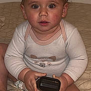 Melina a rejoint le concours — aidez-le/la à gagner de superbes lots ! baby, child, toddler, infant, sitting, indoor, curious, wide_eyes, rosy_cheeks, white_clothing, long_sleeves, quilted_bedspread, holding_object, black_device, cord, closeup, cute, face, person, young