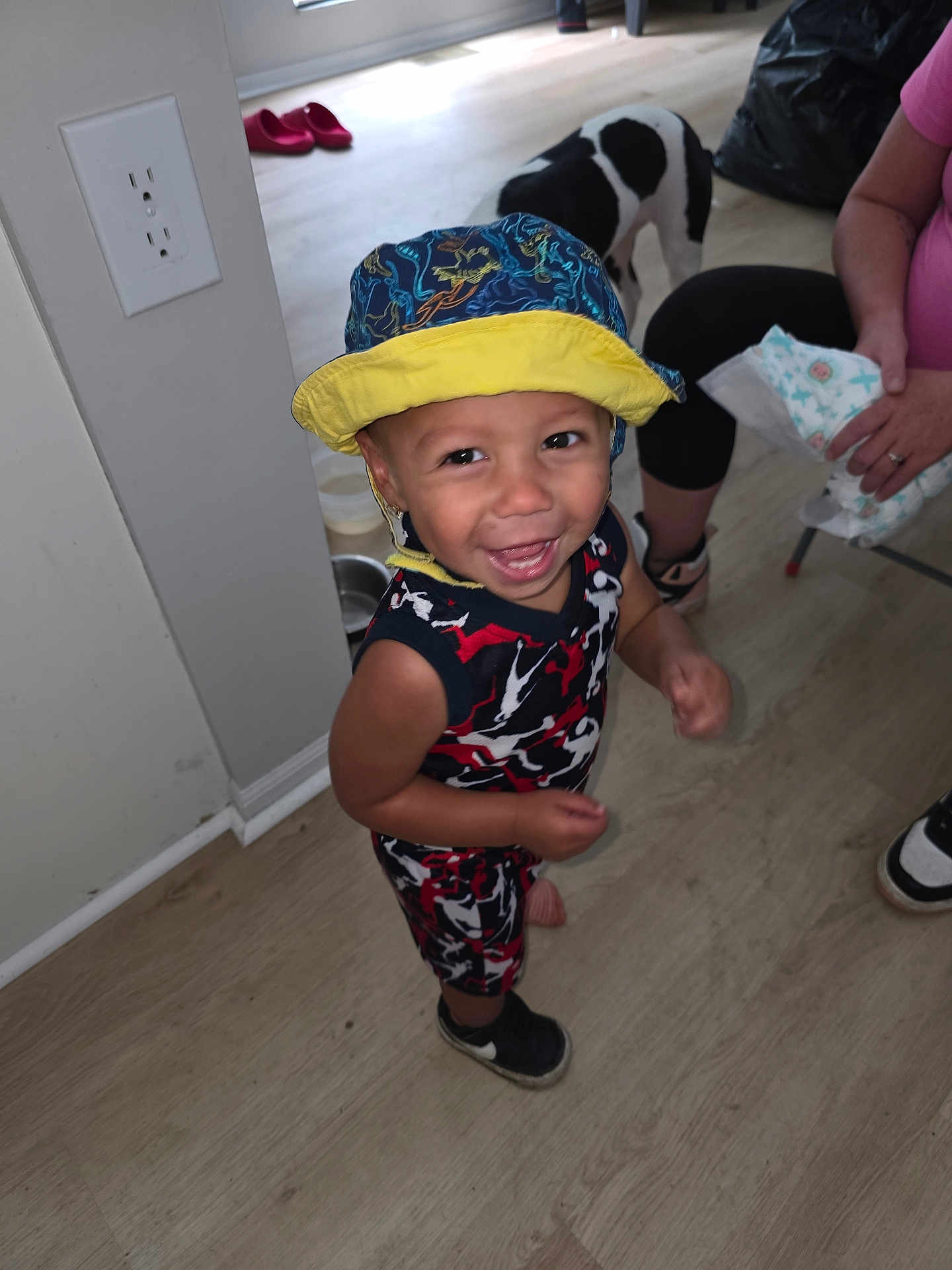 Ryland joined the competition — help win amazing prizes! toddler, child, smiling, hat, bucket_hat, colorful_clothing, indoor, wooden_floor, dog, black_and_white_dog, person, sneakers, happy, playful, floor, wall_outlet, adult, seated, flooring, home