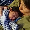 baby, sleeping, striped_clothing, adult, holding, closeup, indoor, blanket, patterned_background, person, face, cozy, resting, parent, child, family, toddler, caring, comfort, warm