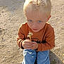 child, blond_hair, blue_eyes, dandelion, sitting, outdoor, casual_clothing, sweater, jeans, nature, sunlight, gravel, curious, young_child, portrait, hands, daylight, innocence, cute, playing