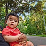 baby, child, person, adult_hand, outdoor, park, trees, greenery, pathway, daylight, nature, casual_clothing, holding, curious_expression, short_sleeve, summer, family, portrait, young_child, relaxation