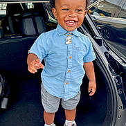 Isaiah is registered to the contest to win money with this photo: toddler, child, smiling, standing, car_trunk, blue_shirt, gray_shorts, sneakers, parking_lot, outdoor, happy, person, curly_hair, jewelry, casual_clothing, daylight, vehicle, urban, cute, fashion