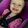 baby, child, smiling, happy, portrait, close_up, car_seat, safety_harness, pink_clothing, purple_background, blue_eyes, teeth, infant, sitting, indoors, vehicle, face, head, photography, cute