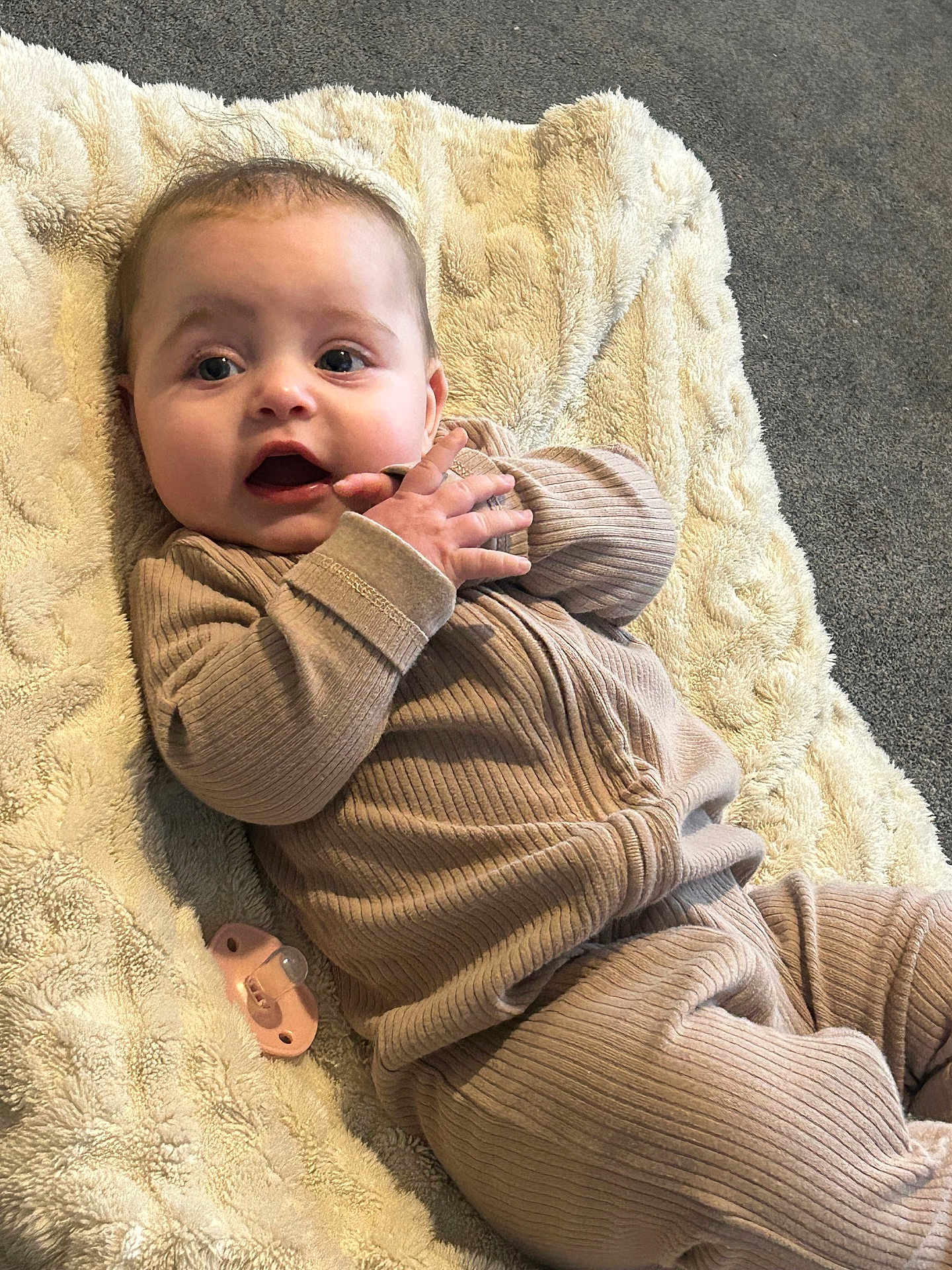 Mina joined the competition — help win amazing prizes! baby, infant, blanket, pacifier, clothing, ribbed_fabric, face, hand, expression, lying_down, floor, carpet, cozy, cute, child, indoor, soft_texture, neutral_colors, portrait, young