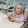 child, toddler, blonde_hair, pink_sweater, stacking_cups, table, classroom, indoor, smiling, playing, person, furniture, toy, happy, young_child, education, seat, background_blur, light, casual_clothing