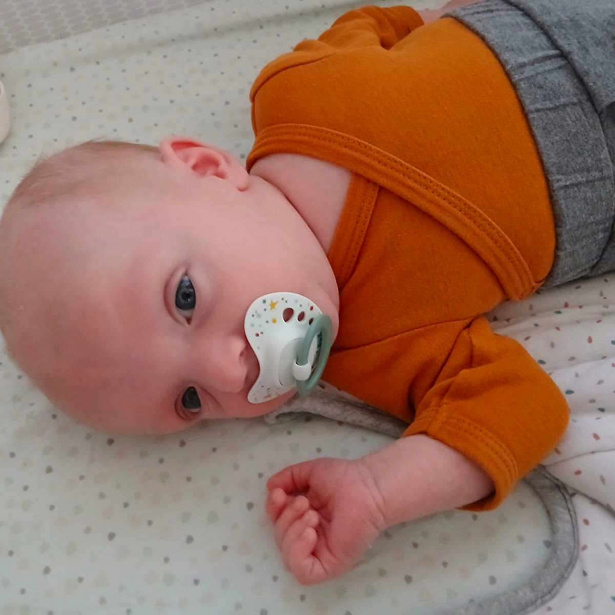 Maylo participe au concours pour gagner de l'argent avec cette photo : baby, blue_eyes, child, cute, face, grey_pants, hand, head, indoors, infant, lying_down, mattress, newborn, orange_clothing, pacifier, patterned_bedding, resting, skin, sleepwear, soft_light