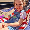 child, girl, smiling, popsicle, summer, outdoor, sunlight, denim_overalls, tie_dye_shirt, chair, american_flag_pattern, pigtails, toy_gun, happy, face, blue_eyes, curly_hair, casual_clothing, playful, daytime