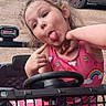 child, girl, playful, silly_face, pink_clothing, rainbow, ice_cream_cone, toy_car, steering_wheel, outdoor, dirt_road, vehicle, curly_hair, red_bow, expression, fun, portrait, person, casual, daylight