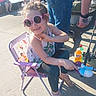 child, girl, sunglasses, toy, water_gun, chair, summer, outdoor, sunny, casual_clothing, sidewalk, waterfront, people, legs, shoes, beverage, tattoo, fishing_rod, playful, smile