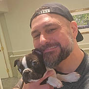 Rollo is registered to the contest to win money with this photo: man, dog, puppy, beard, baseball_cap, tattoo, smile, selfie, indoor, portrait, holding, pet, hand, wall_art, picture_frame, door, carpet, eyes, nose, shirt