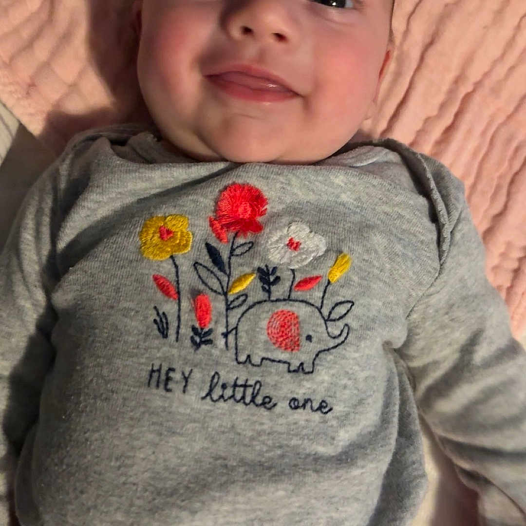 Brooklyn is registered to the contest to win money with this photo: baby, smiling, infant, blanket, clothing, red_pants, grey_shirt, embroidery, elephant, flowers, face, person, child, cute, happy, laying_down, indoors, soft_texture, portrait, closeup