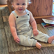 Rowan is registered to the contest to win money with this photo: toddler, child, smiling, sitting, overalls, blonde_hair, indoor, carpet, wooden_furniture, book, cute, happy, baby, person, portrait, floor, eyes, skin, hand, foot
