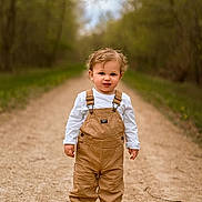 Rowan is registered to the contest to win money with this photo: toddler, child, outdoor, path, forest, trees, overalls, white_shirt, brown_shoes, curly_hair, standing, nature, portrait, daylight, young_child, walking_path, casual_clothing, greenery, dirt_road, innocent