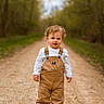 toddler, child, outdoor, path, forest, trees, overalls, white_shirt, brown_shoes, curly_hair, standing, nature, portrait, daylight, young_child, walking_path, casual_clothing, greenery, dirt_road, innocent