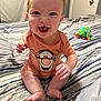 baby, red_hair, smiling, onesie, cartoon_print, bed, striped_bedspread, toy, indoor, child, happy, sitting, person, infant, cute, barefoot, cheerful, skin, hand, face