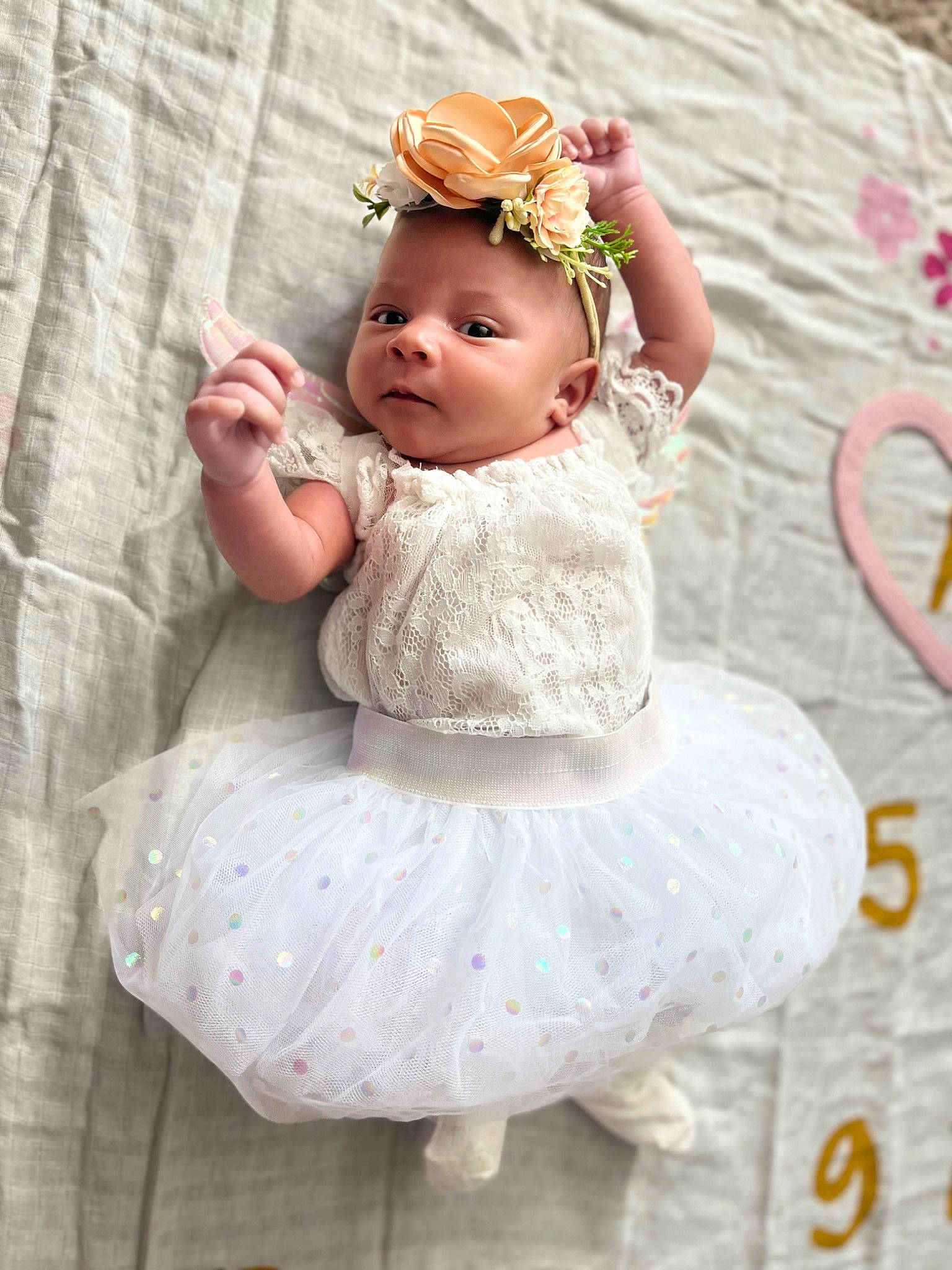 Zariah is registered to the contest to win money with this photo: baby, baby_toddler_clothing, bridal_accessory, child, dress, embellishment, face, flower, hair_accessory, hand, headband, headgear, headpiece, jewellery, pattern, peach, person, petal, pink, sleeve