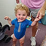 toddler, child, blue_onesie, indoor, person, hand_holding, adult, toy, stroller, carpet, shorts, sneakers, curly_hair, smiling, standing, floor, playful, happy, casual_clothing, skin