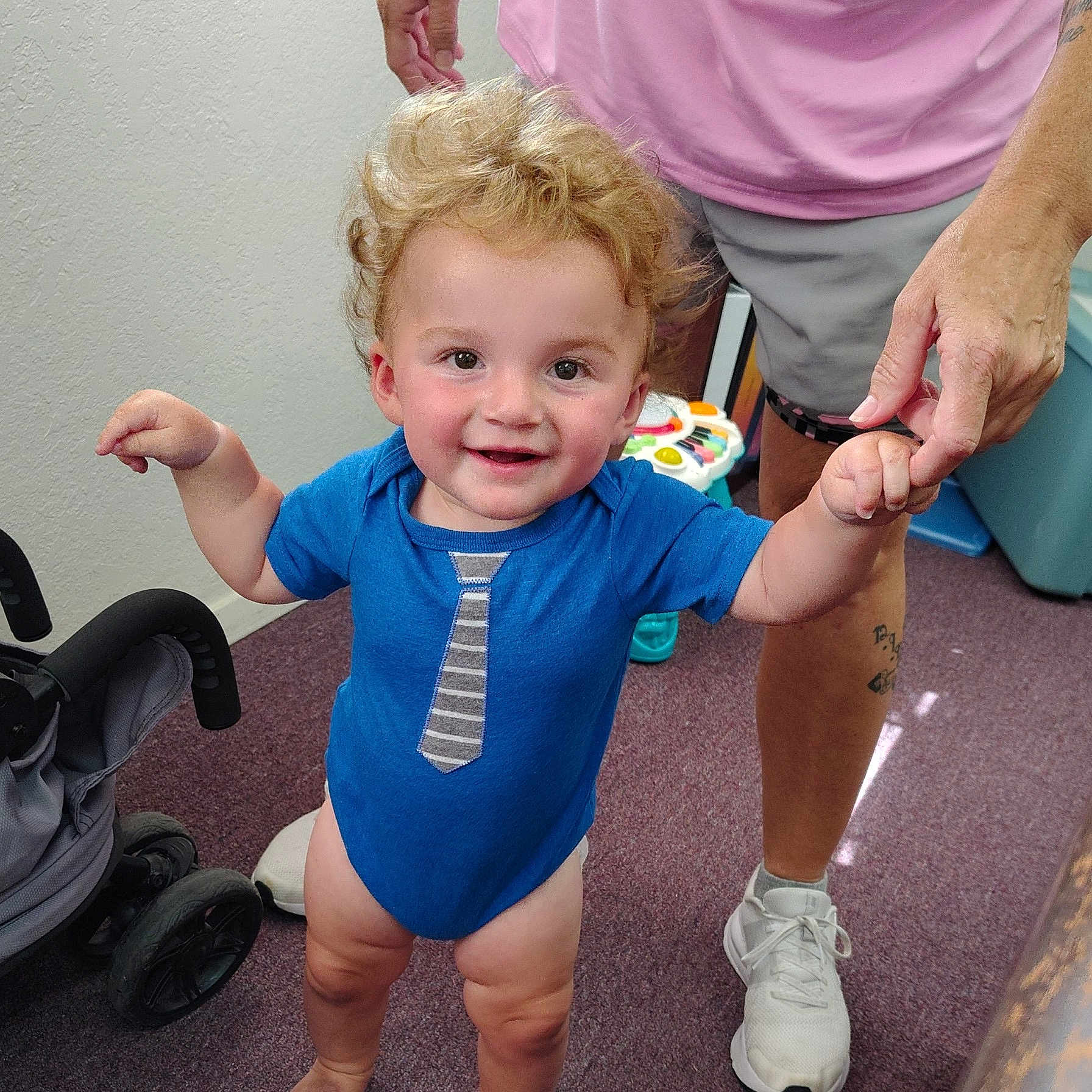 Esiah Michael is registered to the contest to win money with this photo: toddler, child, blue_onesie, indoor, person, hand_holding, adult, toy, stroller, carpet, shorts, sneakers, curly_hair, smiling, standing, floor, playful, happy, casual_clothing, skin