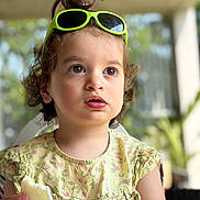 Jaya a rejoint le concours — aidez-le/la à gagner de superbes lots ! toddler, child, portrait, face, eyes, sunglasses, hair, messy_bun, yellow_dress, floral_pattern, hand, food, snack, indoors, blurred_background, curious, expression, person, cute, summer