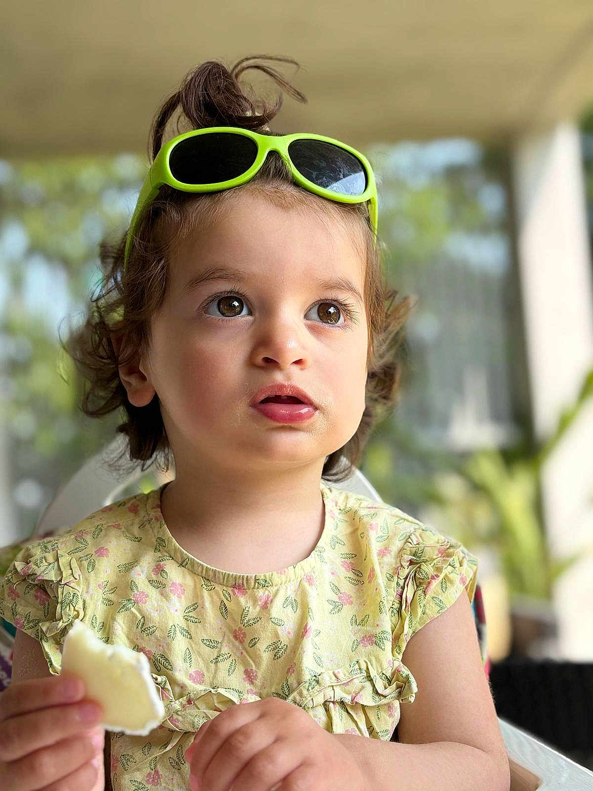 Jaya a rejoint le concours — aidez-le/la à gagner de superbes lots ! toddler, child, portrait, face, eyes, sunglasses, hair, messy_bun, yellow_dress, floral_pattern, hand, food, snack, indoors, blurred_background, curious, expression, person, cute, summer
