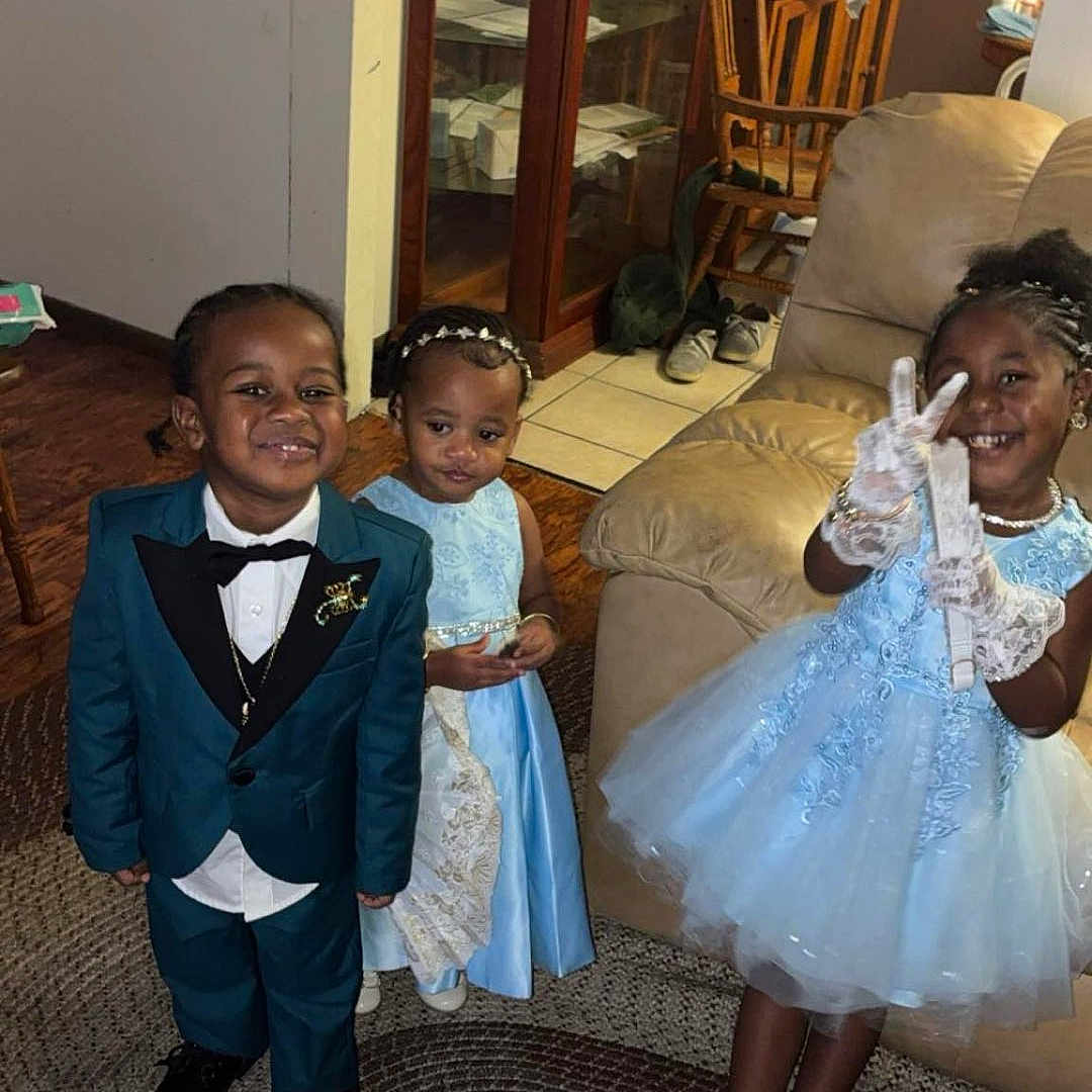 Kharlee is registered to the contest to win money with this photo: cabinet, carpet, child, children, dress, family, floor, footwear, formal_wear, gloves, happy, home, indoor, peace_sign, person, portrait, smiling, sofa, tuxedo, wooden_chair