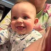 Samson is registered to the contest to win money with this photo: baby, smiling, happy, infant, person, face, child, cute, clothing, onesie, dinosaur_print, indoor, adult, tie_dye_shirt, hand, closeup, portrait, joyful, home, expression