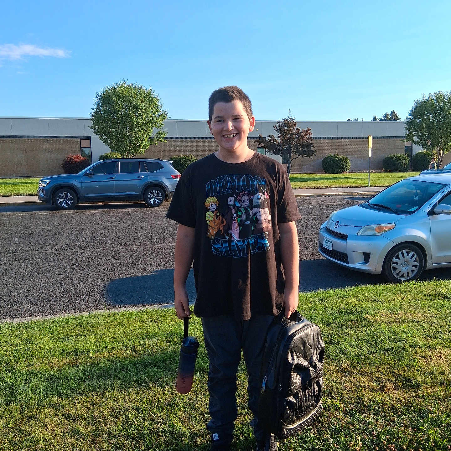Alyrique is registered to the contest to win money with this photo: child, smiling, outdoor, portrait, school, backpack, water_bottle, black_shirt, pants, shoes, grass, sidewalk, street, cars, parking_lot, building, trees, blue_sky, daylight, school_child