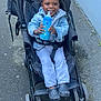 baby_bottle, blue_wall, bottle, casual_clothing, child, curly_hair, daylight, footwear, happy, jacket, outdoor, pants, person, seatbelt, shoes, sidewalk, smile, stroller, toddler, urban