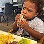 chair, child, clothing, contemplative, curly_hair, daylight, eating, food, meal, meat, outdoor, pasta, person, plate, portrait, restaurant, salad, table, toddler, window